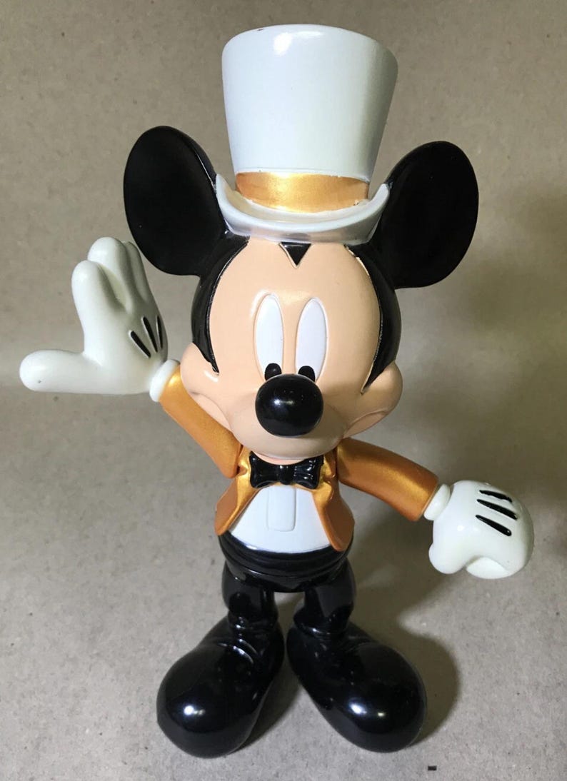 May include: A Mickey Mouse figurine in a formal outfit. The figure wears a white top hat with a gold band, a gold jacket, black pants, and black shoes. Mickey has white gloves and a black bow tie.