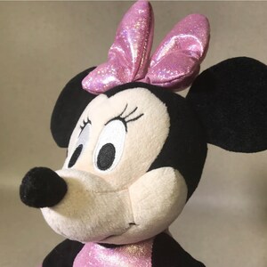 May include: A plush pink and white Minnie Mouse toy with black ears and a pink sparkly bow. The toy has embroidered eyes and a black nose.