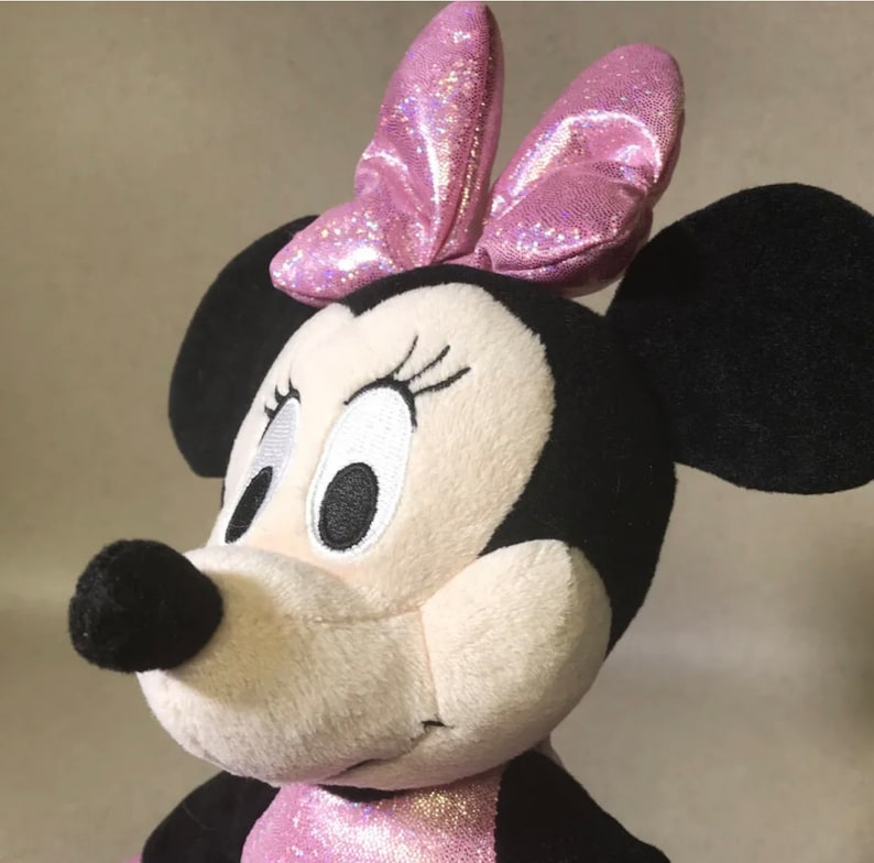 May include: A plush pink and white Minnie Mouse toy with black ears and a pink sparkly bow. The toy has embroidered eyes and a black nose.