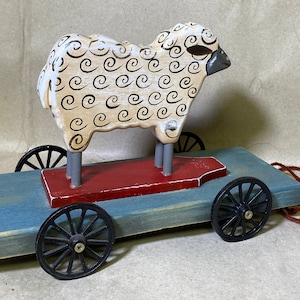 May include: A wooden toy sheep with a black and white swirl pattern on its body, riding on a blue wooden wagon with black wheels. The wagon has a red wooden platform and a pull string with a red ball on the end.