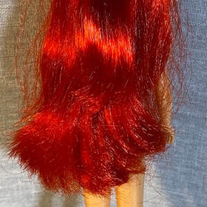 Pu&ograve; includere: Una bambola con i capelli rossi e un fiore tra i capelli. La bambola indossa un costume da bagno e ha un'abbronzatura.