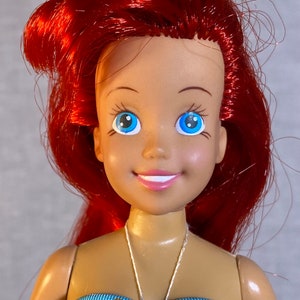 Pu&ograve; includere: Una bambola con i capelli rossi, gli occhi azzurri e un sorriso, che indossa un top di bikini a righe blu, rosa e gialle.