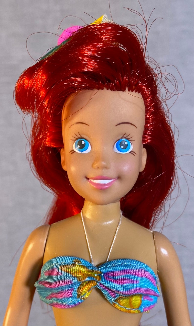Pu&ograve; includere: Una bambola con i capelli rossi, gli occhi azzurri e un sorriso, che indossa un top di bikini a righe blu, rosa e gialle.