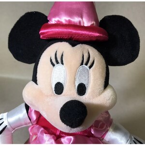 K&ouml;nnte beinhalten: Ein rosa und schwarzes Pl&uuml;schtier von Minnie Mouse, das einen rosa Satin-Hut mit rotem Band tr&auml;gt. Sie hat gro&szlig;e schwarze Augen mit wei&szlig;en Pupillen und lange Wimpern.