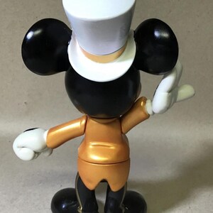 May include: A figurine of a classic cartoon character, viewed from the back. The figure has black ears, a black head, and a white top hat. It wears a gold-colored jacket and black pants with gold accents. White gloves complete the look.