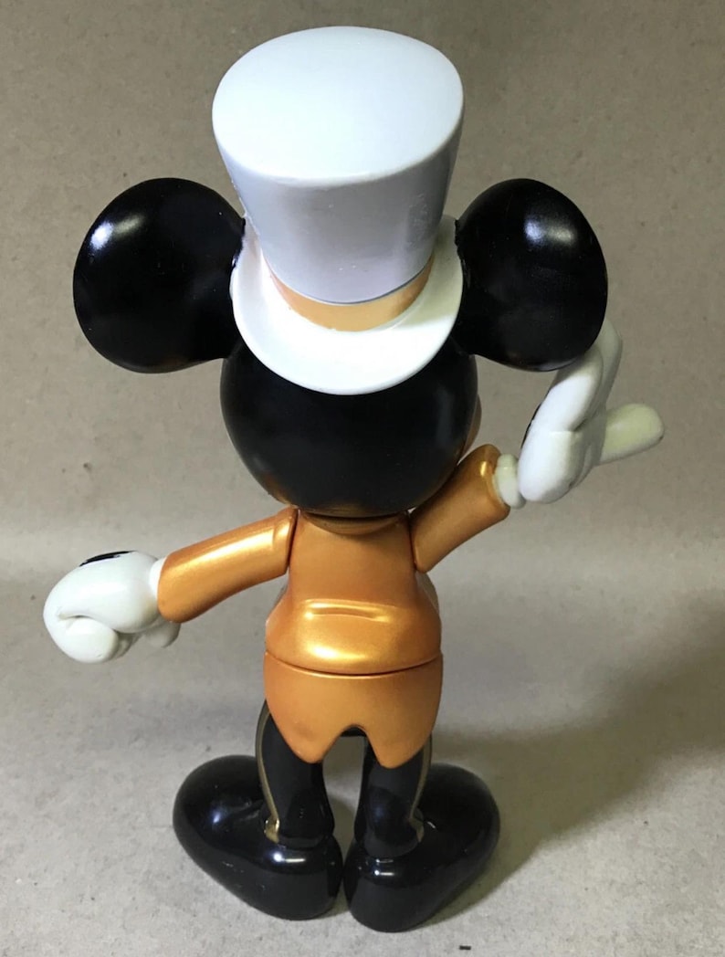May include: A figurine of a classic cartoon character, viewed from the back. The figure has black ears, a black head, and a white top hat. It wears a gold-colored jacket and black pants with gold accents. White gloves complete the look.