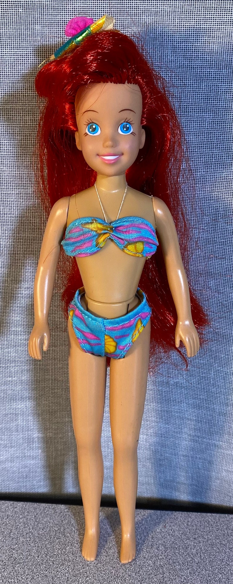 Pu&ograve; includere: Una bambola con i capelli rossi che indossa un bikini a fantasia blu e gialla. La bambola ha gli occhi azzurri e un viso sorridente.