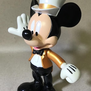 May include: A Mickey Mouse figurine in a formal outfit. The figure wears a white top hat with a gold band, a gold jacket, black pants, and black shoes. Mickey has white gloves and a bow tie, with one hand raised.