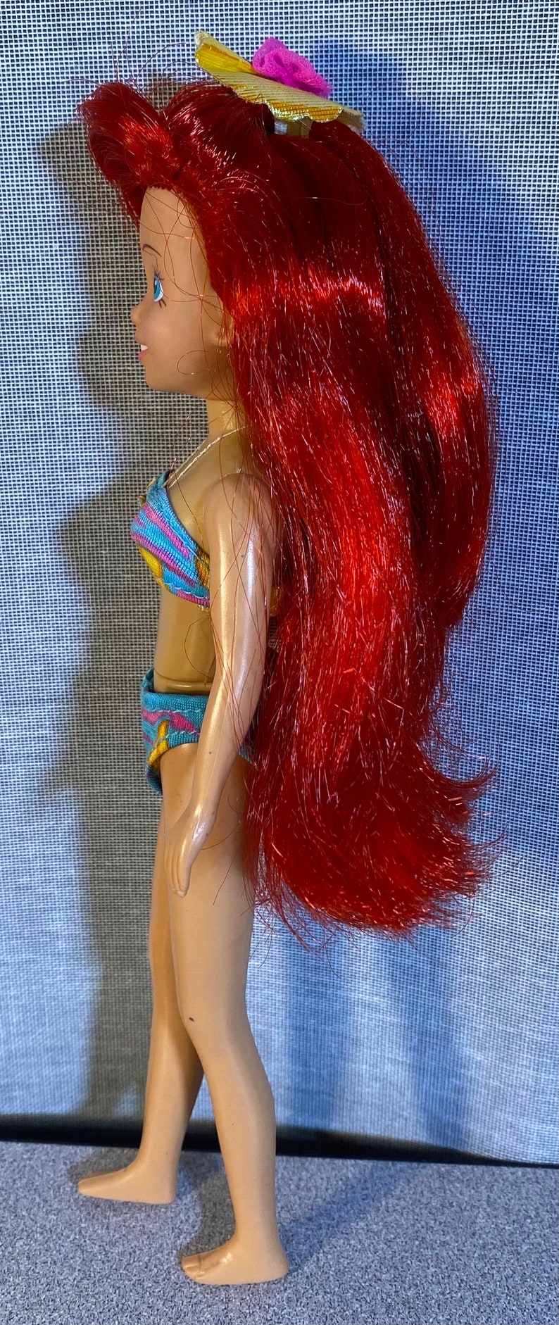 Pu&ograve; includere: Una bambola con i capelli rossi lunghi e fluenti che indossa un costume da bagno blu e giallo e un cappello giallo con un fiore rosa. La bambola &egrave; in piedi su un tappeto grigio.