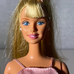 May include: A blonde doll with blue eyes wearing a pink satin dress and a pink bow in her hair.