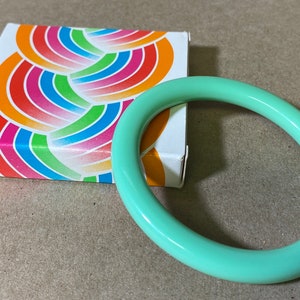 May include: A light green plastic bangle bracelet. The bracelet is thick and has a smooth, glossy finish. It is sitting on a brown surface next to a small box with a colorful rainbow pattern.
