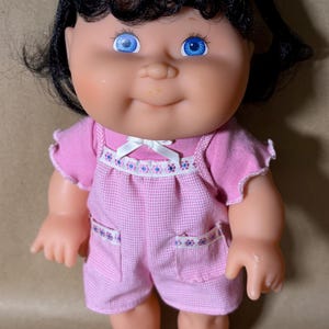 May include: A doll with black hair styled in two pigtails with pink bows, bright blue eyes, and a light skin tone. The doll wears a pink short-sleeved top and pink gingham overalls featuring a white ribbon bow and floral trim on the pockets.