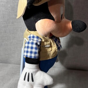 May include: A Mickey Mouse plush toy wearing a tan safari hat with a red band, a tan vest over a blue and white checkered shirt, and blue pants. The toy has white gloves and an orange shoe.