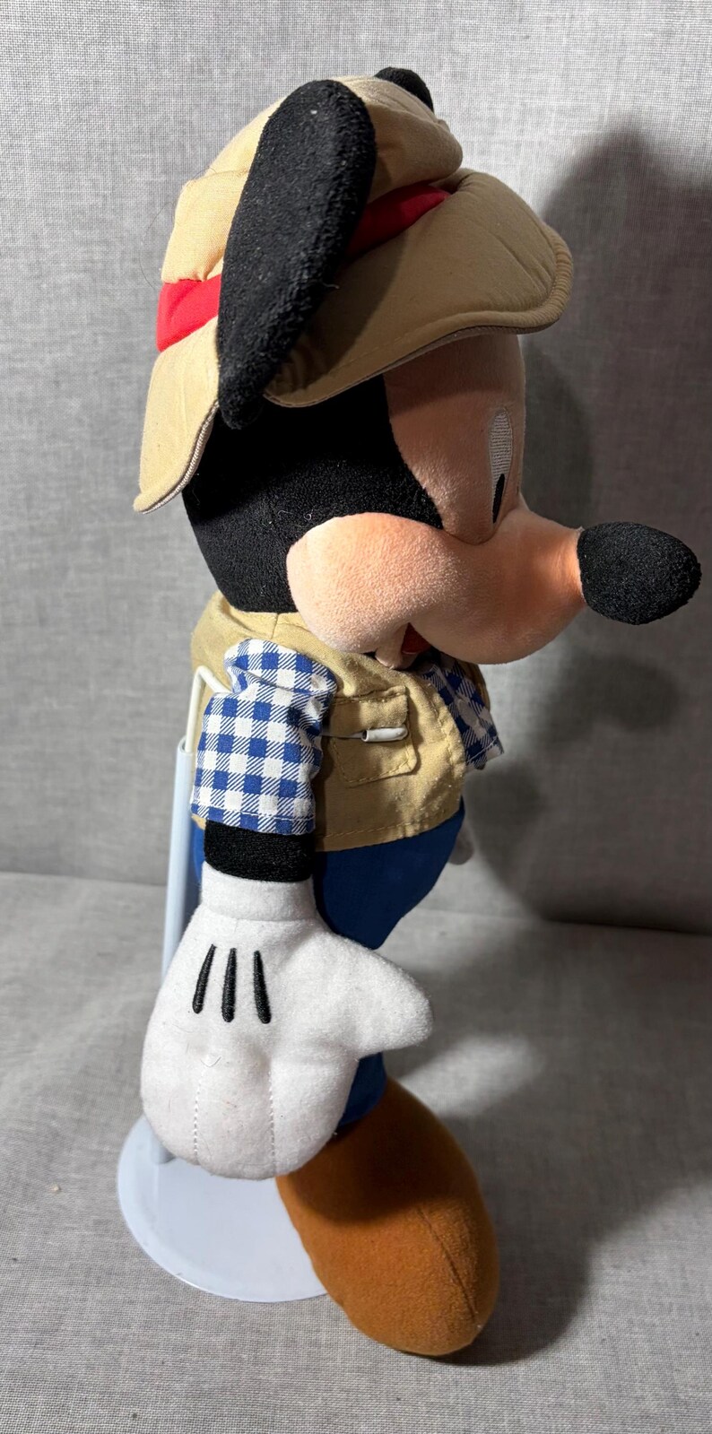 May include: A Mickey Mouse plush toy wearing a tan safari hat with a red band, a tan vest over a blue and white checkered shirt, and blue pants. The toy has white gloves and an orange shoe.
