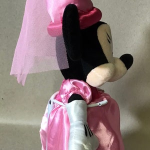 K&ouml;nnte beinhalten: Ein rosa Pl&uuml;sch-Minnie Mouse, verkleidet als Prinzessin. Sie tr&auml;gt ein rosa Kleid, eine rosa Krone und einen wei&szlig;en Schleier. Sie steht auf einem wei&szlig;en Sockel.