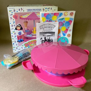 May include: A pink plastic soda fountain playset with a spinning carousel top and two pink seats. The set includes two dolls, a table, and a variety of accessories. The box features the text "Bouncin' Kids" and "Soda Fountain".