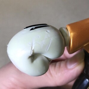 May include: Close-up of a light green figurine head with black eye markings. The figurine is connected to a gold and black body. The head has visible imperfections.