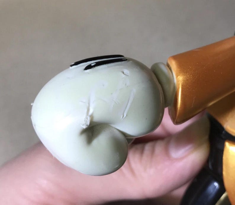May include: Close-up of a light green figurine head with black eye markings. The figurine is connected to a gold and black body. The head has visible imperfections.