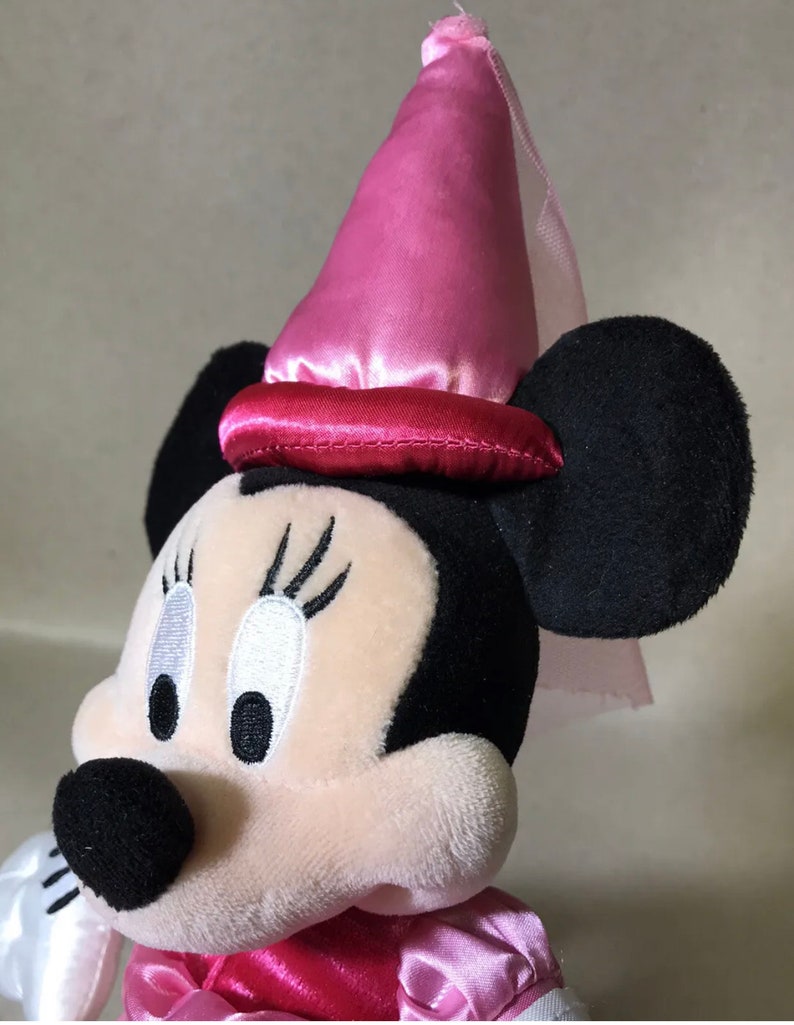 K&ouml;nnte beinhalten: Ein rosa-schwarzes Pl&uuml;schtier von Minnie Mouse, das einen rosa Satin-Hut mit Schleier tr&auml;gt. Der Hut hat ein rotes Satinband um die Basis.