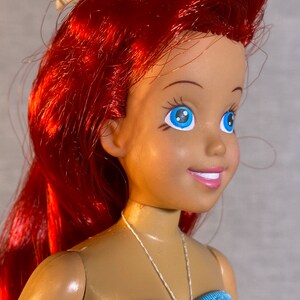 Pu&ograve; includere: Una bambola con i capelli rossi e gli occhi azzurri che indossa un top di bikini a righe blu, rosa e gialle. La bambola ha un fiocco rosa e verde sulla testa.