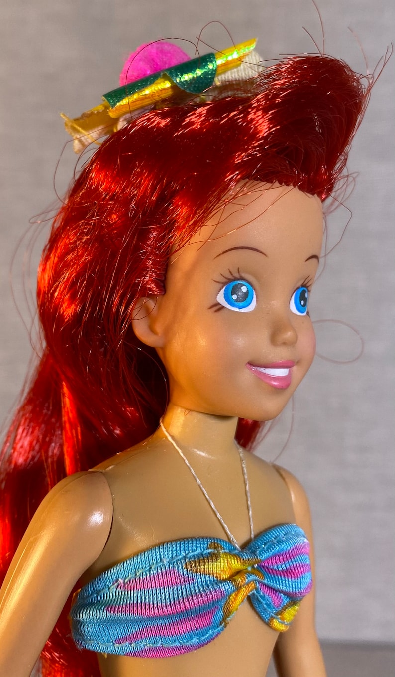 Pu&ograve; includere: Una bambola con i capelli rossi e gli occhi azzurri che indossa un top di bikini a righe blu, rosa e gialle. La bambola ha un fiocco rosa e verde sulla testa.
