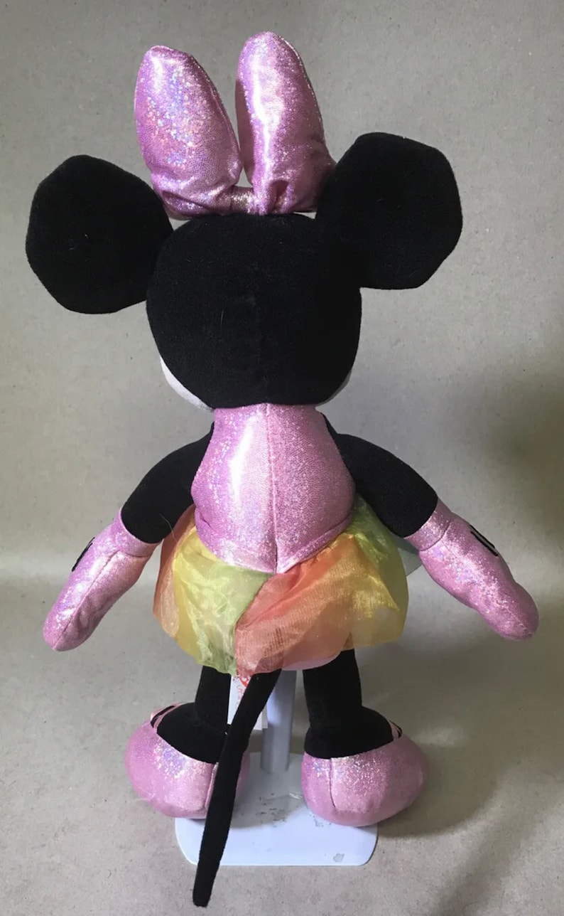 May include: A pink and black plush Minnie Mouse toy wearing a pink sparkly dress and a pink bow. The dress has a rainbow tulle skirt.