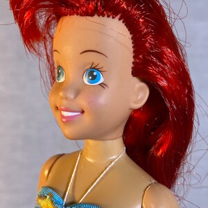 Pu&ograve; includere: Una bambola con i capelli rossi e gli occhi azzurri che indossa un top di bikini a fantasia blu, gialla e rosa. La bambola ha una collana di corda bianca.
