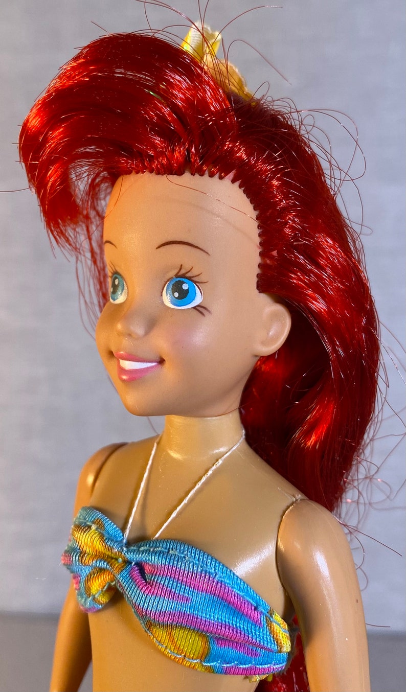 Pu&ograve; includere: Una bambola con i capelli rossi e gli occhi azzurri che indossa un top di bikini a fantasia blu, gialla e rosa. La bambola ha una collana di corda bianca.