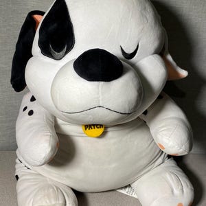 May include: White and black plush Dalmatian dog with a yellow tag that says "Patch". The dog is sitting with its head tilted and one eye closed.