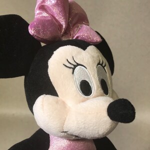 May include: A plush pink and black Minnie Mouse toy with a pink sparkly bow on her head. She is wearing a pink sparkly dress.