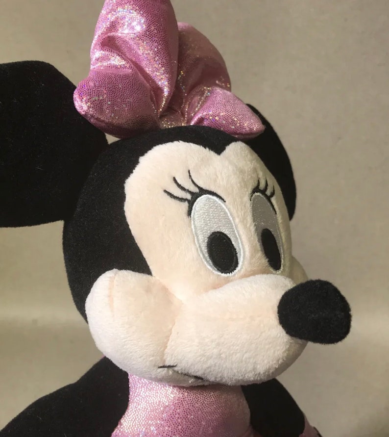 May include: A plush pink and black Minnie Mouse toy with a pink sparkly bow on her head. She is wearing a pink sparkly dress.