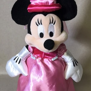 K&ouml;nnte beinhalten: Ein rosa Pl&uuml;sch-Minnie Mouse, verkleidet als Prinzessin. Sie tr&auml;gt ein rosa Kleid mit wei&szlig;em Satinbesatz, eine rosa Krone mit wei&szlig;em Schleier und rosa Schuhe.