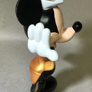 May include: A figurine of a cartoon mouse wearing a white top hat, black ears, and a black body with gold accents. The mouse has white gloves and a black nose. The figurine is in a side profile pose.