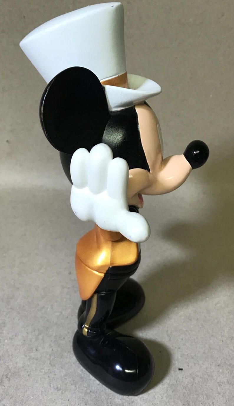 May include: A figurine of a cartoon mouse wearing a white top hat, black ears, and a black body with gold accents. The mouse has white gloves and a black nose. The figurine is in a side profile pose.