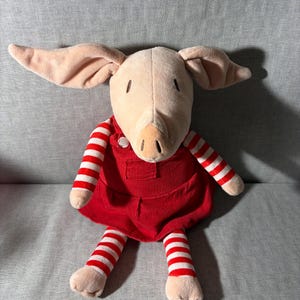 May include: A plush pig doll with a light pink face, ears, and feet. The pig is wearing a red corduroy dress and red and white striped leggings. The doll is sitting on a gray surface.