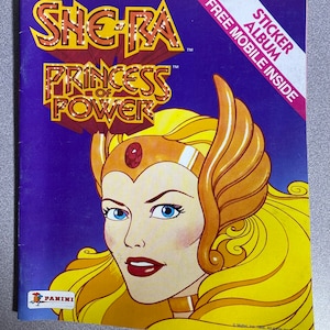 Może przedstawiać: Okładka albumu z naklejkami She-Ra: Princess of Power w stylu vintage. Okładka przedstawia portret She-Ra z blond włosami i złotą koroną na fioletowym tle. Album zawiera tekst "STICKER ALBUM" i "FREE MOBILE INSIDE".