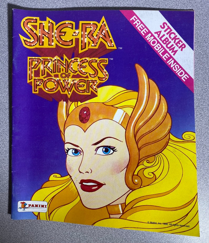 Może przedstawiać: Okładka albumu z naklejkami She-Ra: Princess of Power w stylu vintage. Okładka przedstawia portret She-Ra z blond włosami i złotą koroną na fioletowym tle. Album zawiera tekst "STICKER ALBUM" i "FREE MOBILE INSIDE".