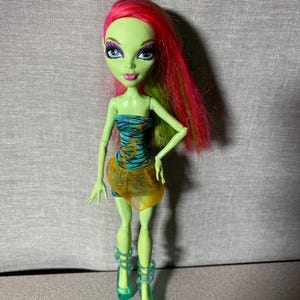 Peut inclure: Poupée Monster High à la peau verte, aux cheveux roses et verts et aux yeux bleus. Elle porte un haut sans bretelles turquoise et orange, une jupe jaune et des talons verts translucides. La poupée est posée sur un fond gris.