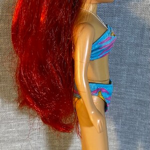 Pu&ograve; includere: Una bambola con i capelli rossi che indossa un costume da bagno blu e rosa. La bambola ha un fermaglio per capelli a forma di fiore nei capelli.