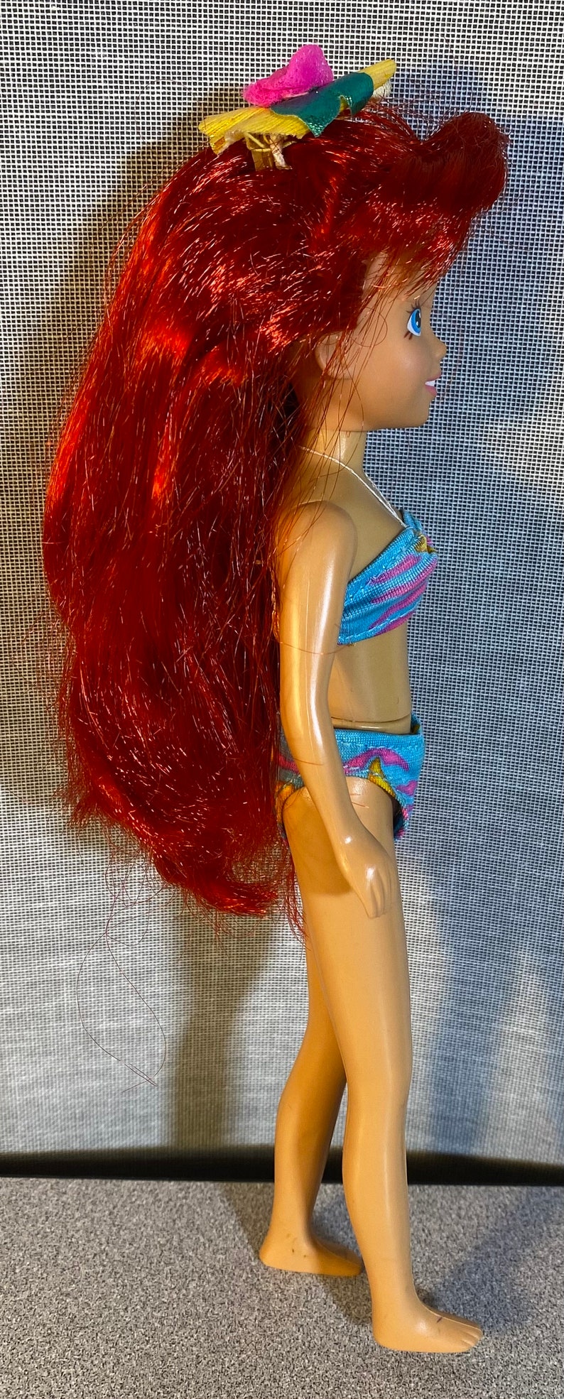Pu&ograve; includere: Una bambola con i capelli rossi che indossa un costume da bagno blu e rosa. La bambola ha un fermaglio per capelli a forma di fiore nei capelli.