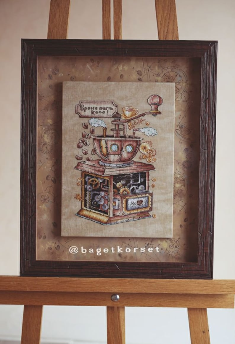 Op de afbeelding: Een ingelijst kruissteekkunstwerk dat een stoom-aangedreven koffiemolen afbeeldt met de tekst "Время пить кофе!" (Tijd om koffie te drinken!) in het Russisch. Het kunstwerk is ingelijst in een donkerbruin houten lijst en wordt op een ezel tentoongesteld.