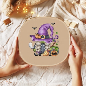 Pu&ograve; includere: Un cerchio da ricamo a punto croce con un gnomo di Halloween che indossa un cappello da strega viola e tiene una scopa. Lo gnomo &egrave; circondato da zucche, un calderone e un ragno.