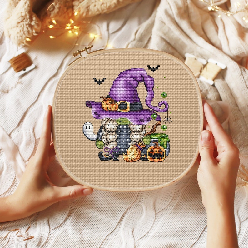 Pu&ograve; includere: Un cerchio da ricamo a punto croce con un gnomo di Halloween che indossa un cappello da strega viola e tiene una scopa. Lo gnomo &egrave; circondato da zucche, un calderone e un ragno.