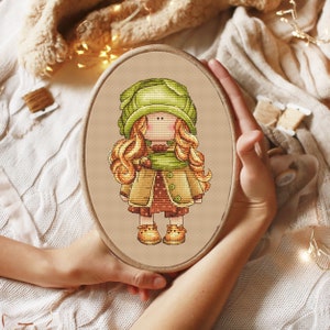 May include: A cross-stitch embroidery of a girl wearing a green hat, brown coat, and brown boots. The girl has long blonde hair and is holding a small brown object in her hand.