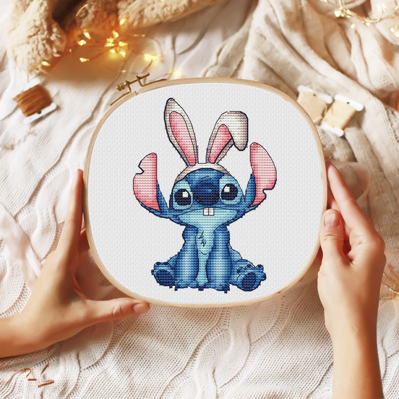 May include: A blue Stitch character from the Disney movie Lilo & Stitch wearing bunny ears. The character is sitting down with its arms crossed and a big smile on its face. The image is a cross-stitch pattern.