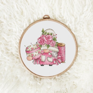 May include: A cross-stitch pattern of a gnome wearing a pink dress and a hat with pink roses. The gnome is holding a pincushion and a spool of thread.