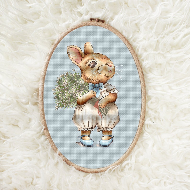 以下が含まれることがあります： 白い花束を持った茶色のウサギのクロスステッチ刺繍。ウサギは青いリボンと青い靴が付いた白いドレスを着ています。刺繍は薄い茶色の楕円形の枠に囲まれています。