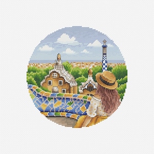 May include: A circular cross-stitch embroidery depicting a person with long brown hair wearing a straw hat, looking out over a park with colorful mosaic walls, brown buildings with unique rooflines, green trees, and a blue sky with white clouds.