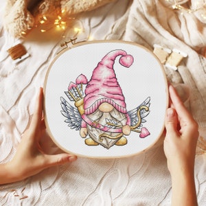 Puede incluir: Un patrón de punto de cruz de un gnomo rosa con alas que sostiene una flecha y corazones. El gnomo lleva un gorro rosa con un corazón en la parte superior. El patrón está sobre un fondo blanco.