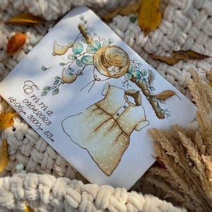 Peut inclure: Un panneau en tissu blanc avec une broderie au point de croix repr&eacute;sentant une robe de b&eacute;b&eacute; jaune, un chapeau de paille et des feuillages. Le texte indique Emma, 09.04.2023, 18:06pm, 3500g, 53cm. Broderie aux tons jaunes, verts et bruns.